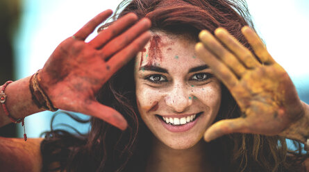 Young woman holding paint covered hands up to her face, one red and one yellow, smiling, Framing her face. - MINF08614