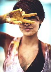 Junge Frau, die sich zwei mit Farbe verschmierte Finger vor das Gesicht hält und in die Kamera schaut; Kleidung und Haut mit Farbe bedeckt. - MINF08615