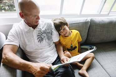 High angle view of bald man and young boy with brown hair sitting side by side on a sofa, reading. - MINF08616