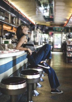 Side view of woman wearing jeans and sleeveless top leaning against a bar counter in a restaurant, looking up. - MINF08619