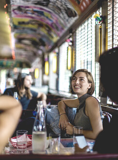 Smiling woman sitting in a booth in a diner looking up and talking to friends. - MINF08623