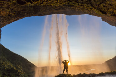 Rückansicht eines Mannes, der bei Sonnenuntergang unter einem natürlichen Bogen hinter einem Wasserfall steht. - MINF08735