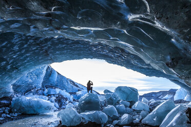 Rückansicht der Silhouette einer Person, die auf einem Eisfelsen am Eingang einer Gletschereishöhle steht. - MINF08737