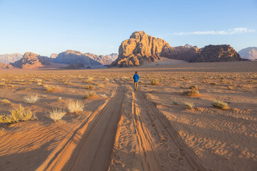 Rear view of man walking in desert landscape with tire tracks leading to distant mountain. - MINF08741