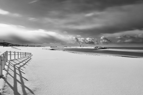 Ein Zaun, der im Winter an einem schneebedeckten Strand in der Nähe des Meeres verläuft., lizenzfreies Stockfoto
