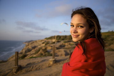 Female in her early 20's wrapped in a red blanket smiles at the camera while she watches the paragliders at sunset over the ocean. - AURF00626