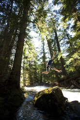 A young man hiking jumps off a rock next to a river in the forest. - AURF00719