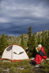 Erwachsene Frau trinkt Kaffee in einem Schlafsack in der Nähe ihres Zeltes beim Rucksackcamping. - AURF00784