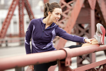 Eine sportliche Frau in einer lila Jacke, die sich auf einer Brücke in Portland, Oregon, streckt. - AURF00796