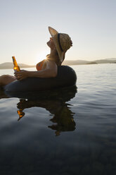 Eine junge Frau genießt die letzten Sonnenstrahlen des Tages, während sie mit einem Bier in einem Schlauchboot auf einem See schwimmt. - AURF00812