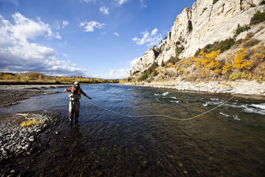 A athletic man fly fishing stands in a river with the fall colors and snowy mountains behind him. - AURF01039