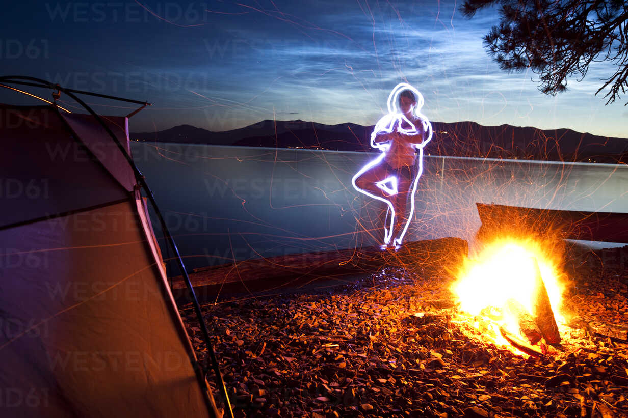 A light painting of a woman doing yoga next to her tent and campfire on a  camping trip along the shores of a lake in Idaho. stock photo