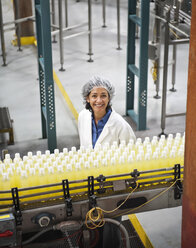 Porträt einer afroamerikanischen Arbeiterin, die ein Kopfnetz trägt und neben einem Förderband für Wasser mit Zitronengeschmack in einer Abfüllanlage steht. - MINF08929