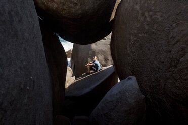 Ein Mann sitzt im Sonnenlicht vor einer Höhle aus riesigen Felsbrocken, die die Südspitze von Virgin Gorda, The Baths National Park, British Virgin Islands, bedecken. - AURF01154