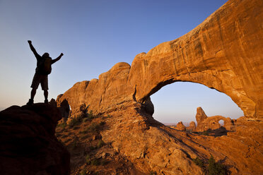 A silhouetted hiker raises his arms beside an arch in Arches National Park, Utah. - AURF01200