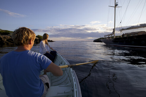 Ein junger Mann und ein Mädchen rudern in einem Peapod-Schlauchboot bei Sonnenuntergang zu einer klassischen Yacht, die an der Küste von Maine vor Anker liegt. - AURF01253