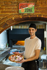 Porträt eines lächelnden Pizzabäckers, der eine Pizza in der Küche hält - AFVF01449
