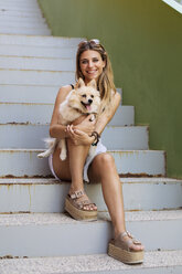 Lächelnde junge Frau sitzt auf einer Treppe und hält ihren Hund - ACPF00256