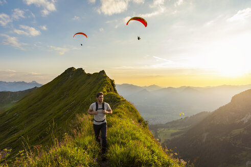 Germany, Bavaria, Oberstdorf, man on a hike in the mountains at sunset with paraglider in background - DIGF04988