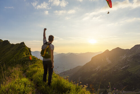 Deutschland, Bayern, Oberstdorf, Mann auf einer Wanderung in den Bergen bei Sonnenuntergang mit Paraglider im Hintergrund - DIGF04990