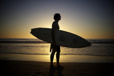 Silhouette eines Surfers, der ein Surfbrett am Strand während des Sonnenuntergangs hält. - AURF01495