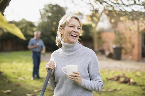 Glückliche reife Frau trinkt Kaffee und harken Herbstblätter im Hinterhof, lizenzfreies Stockfoto