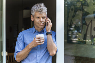Porträt eines reifen Mannes am Telefon, der sich mit einem Kaffeebecher an eine Tür lehnt - TCF05651