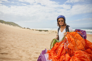 Porträt lächelnder männlicher Gleitschirmflieger mit Fallschirm am Strand - CAIF21693