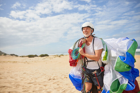 Lächelnder männlicher Gleitschirmflieger mit Ausrüstung und Fallschirm am sonnigen Strand - CAIF21705