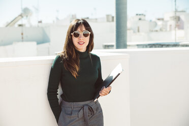 Businesswoman with sunglasses standing on rooftop, holding laptop - SUF00543