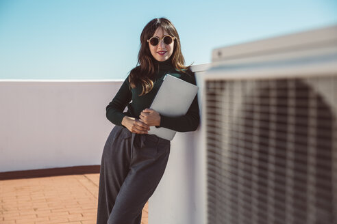 Businesswoman with sunglasses standing on rooftop, holding laptop - SUF00544