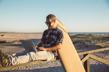 Mann am Strand sitzend, mit Laptop, mit Surfbrett an Zaun gelehnt - SUF00561
