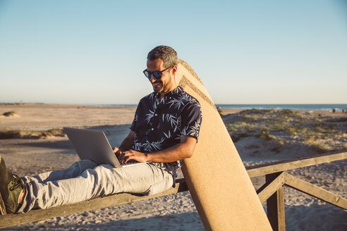 Man sitting at the beach, using laptop, with surfboard leaning on fence - SUF00561