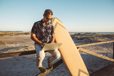 Mann am Strand sitzend, mit Laptop, mit Surfbrett an Zaun gelehnt - SUF00562