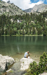 Spanien, Vater und Tochter sitzen auf einem Felsen an einem Bergsee und füttern Enten - GEMF02349