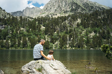 Spanien, Vater und Tochter sitzen auf einem Felsen an einem Bergsee und füttern Enten - GEMF02350