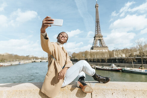 Frankreich, Paris, Frau sitzt auf Brücke über die Seine mit dem Eiffelturm im Hintergrund und macht ein Selfie - KIJF02010