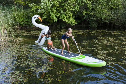 Zwei Mädchen in einem Teich mit aufblasbarem Poolspielzeug in Schwanenform und SUP-Board, lizenzfreies Stockfoto