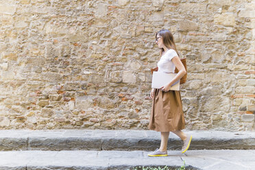 Woman walking along a stone wall carrying laptop - FBAF00005