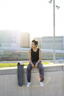 Junger Mann sitzt bei Sonnenuntergang auf einer Stadtmauer neben einem Skateboard - AFVF01483
