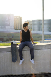 Junger Mann sitzt bei Sonnenuntergang auf einer Stadtmauer neben einem Skateboard - AFVF01484