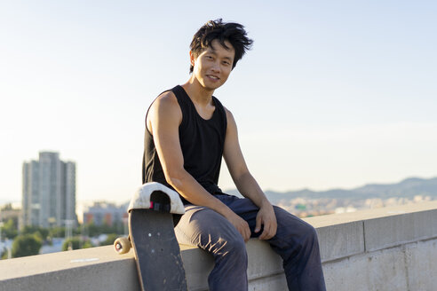 Porträt eines lächelnden jungen Mannes, der neben einem Skateboard an der Wand sitzt - AFVF01486