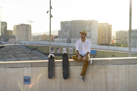 Junger Mann sitzt bei Sonnenuntergang auf einer Stadtmauer neben einem Skateboard, lizenzfreies Stockfoto