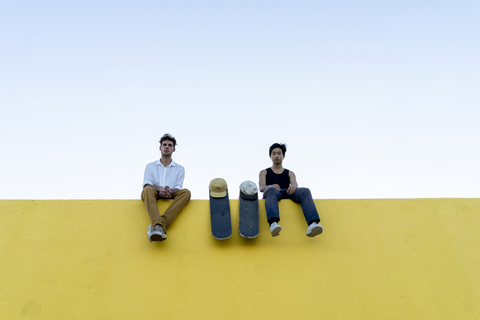 Zwei junge Männer mit Skateboards sitzen auf einer hohen gelben Mauer, lizenzfreies Stockfoto
