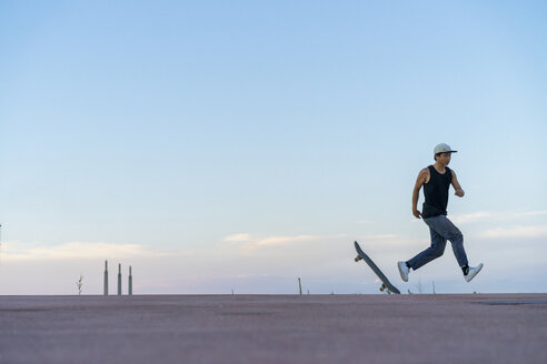 Junger Mann läuft neben Skateboard auf einer Fahrbahn in der Abenddämmerung - AFVF01515