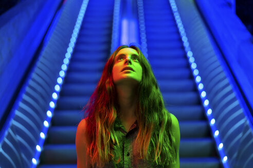 Porträt einer beleuchteten jungen Frau, die vor einer blau beleuchteten Rolltreppe steht und nach oben schaut - AFVF01524