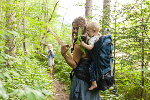 Frau wandert im Wald und zeigt einem kleinen Jungen im Rucksack ein großes Blatt, lizenzfreies Stockfoto