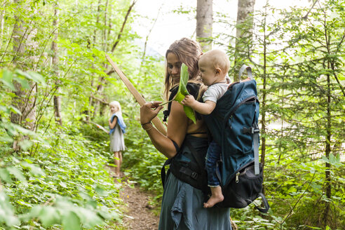 Woman hiking in the woods showing large leaf to baby boy in backpack - TCF05766