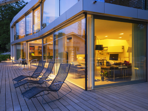 Schweiz, drei Liegestühle auf der Terrasse einer beleuchteten modernen Villa in der Abenddämmerung, lizenzfreies Stockfoto