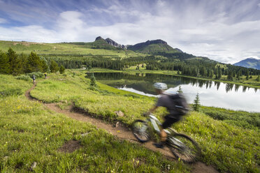 Ein Mountainbiker fährt unscharf durch eine grüne Berglandschaft mit einem See im Hintergrund. - AURF02006